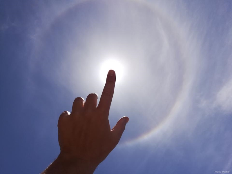 sun on a blue sky with a ring of light around it, and an outstretched finger overlapping the sun itself