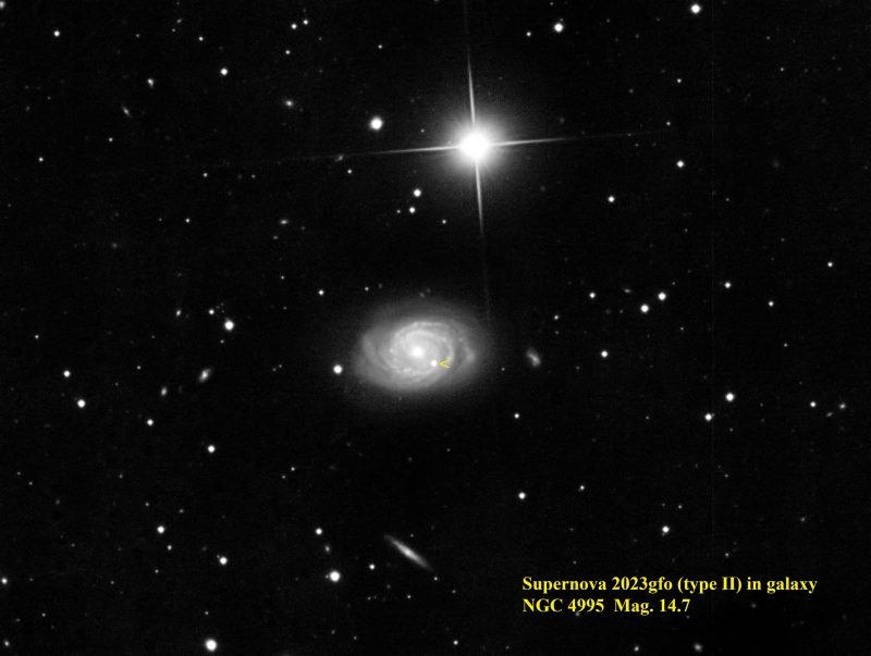 A grayish spiral with a bright star and fainter foreground stars, plus a small yellow arrowhead.