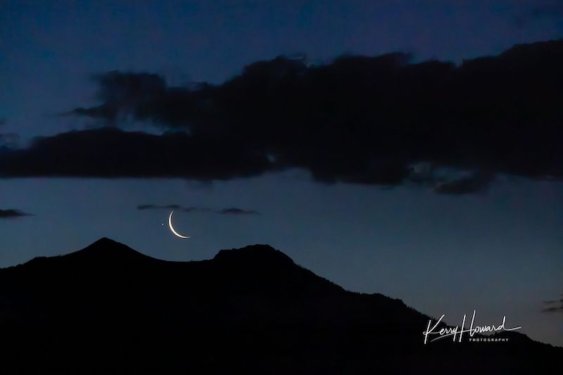 Jupiter near waning crescent moon in a dark blue and cloudy sky with hills in foreground.