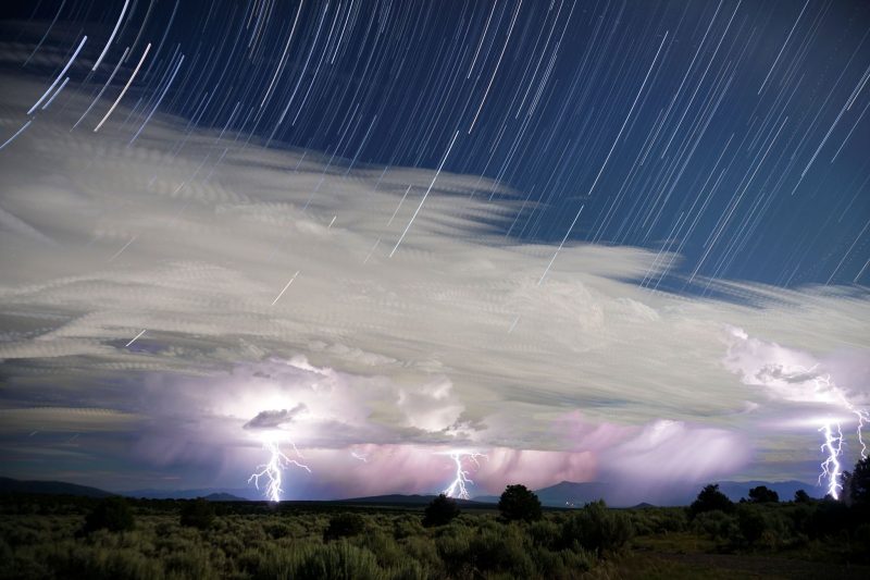Many thin white parallel lines in dark sky above white clouds with lightning bolts reaching the ground below.