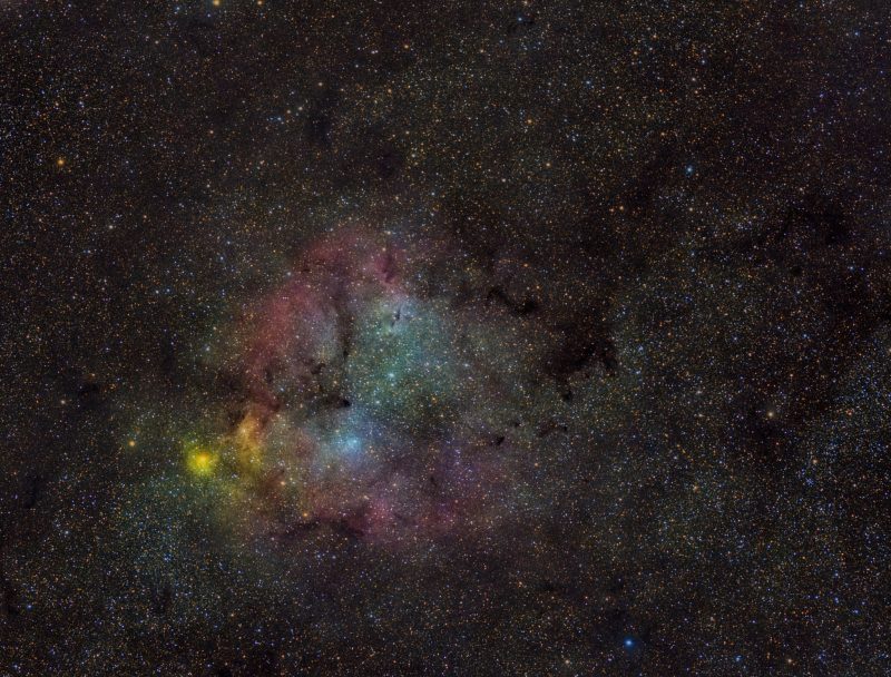 Large area of yellow, red and blue nebulosity with dark lanes, over a background of faint stars.