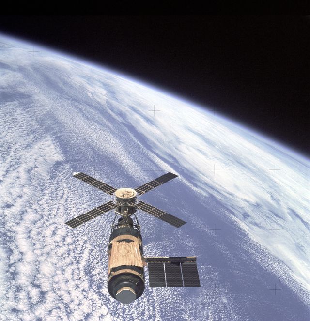 Space station with 4 blades like a helicopter, above cloudtops, with curve of Earth visible.