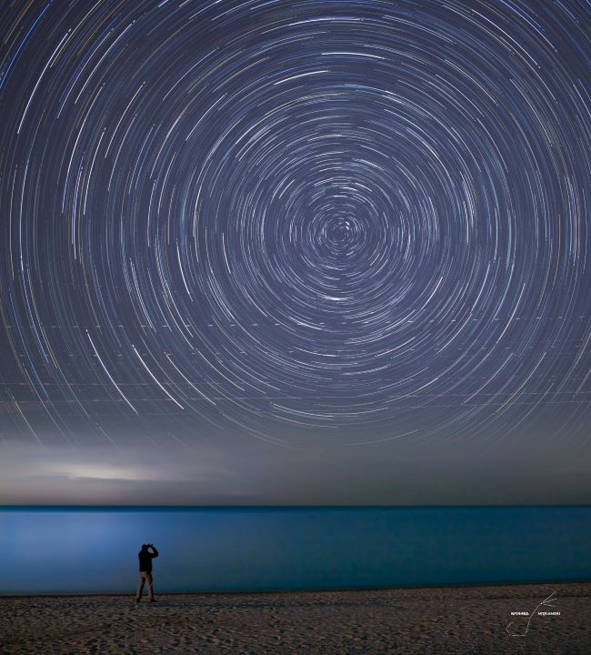 A man on the beach at night photographing the sky, with a circle of concentric dashes made from starlight overhead.