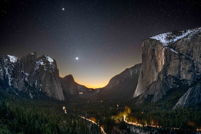 Stargazing and the national parks: Night sky over a valley covered in trees. There are huge cliffs on both sides, lights of cars below and a lit-up waterfall on the right.