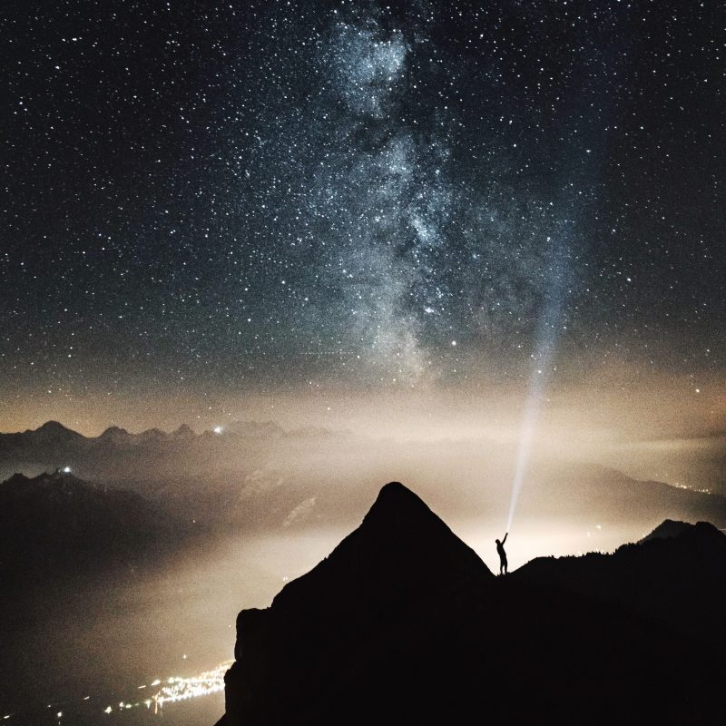 Aliens: A person on a dark hill above a bright town shines a flashlight at the Milky Way.
