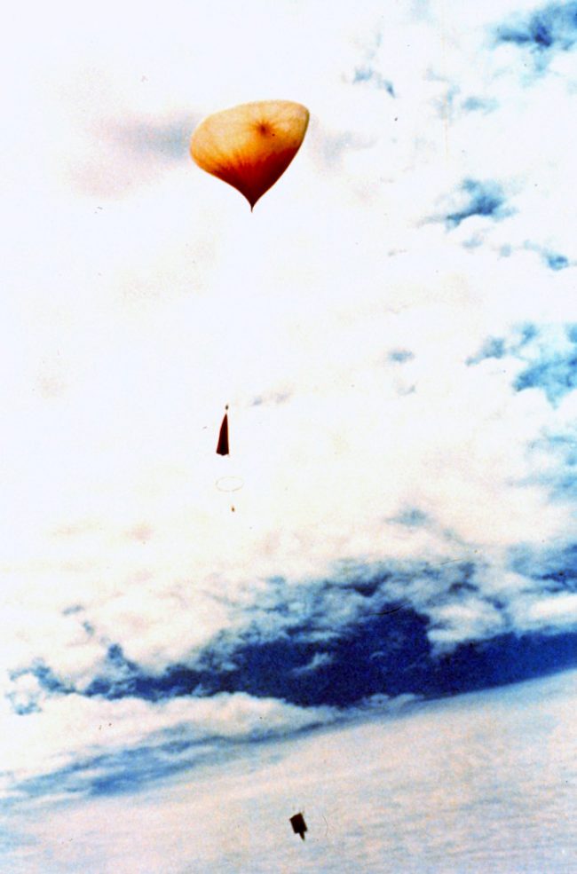 Orangish squished balloon shape floating upward into clouds with stuff dangling below.