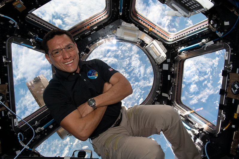 Human body: Man with dark hair and steel-rimmed glasses floating in front of windows on space station.
