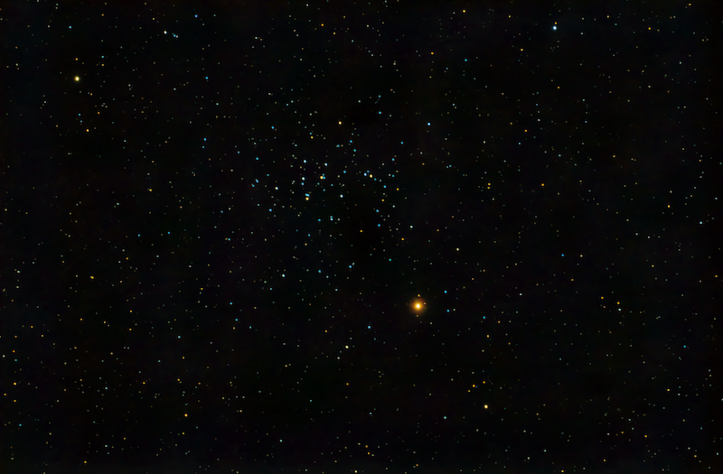 A starry sky with a group of dozens of stars is relatively close together with the glowing reddish planet Mars nearby.