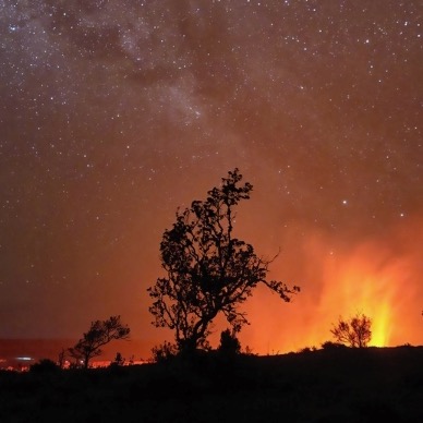 Fiery plumes of a volcano with trees in the foreground.