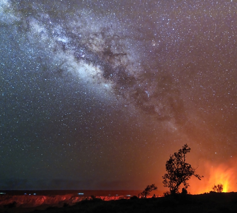 Kilauea: Very bright clouds of the milky way arc from top left to bottom right, where you can see the fiery plumes of a volcano.