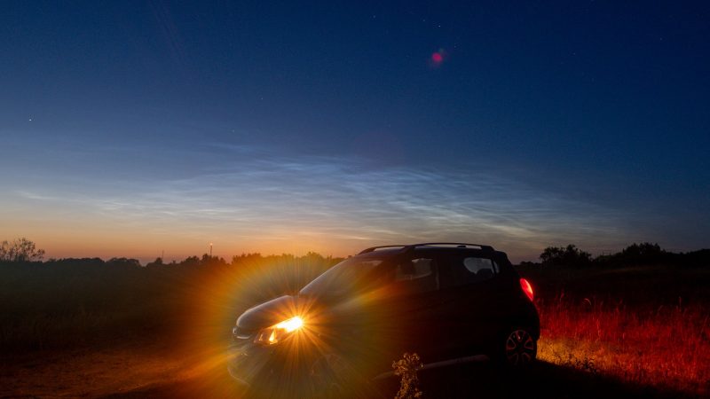 Noctilucent clouds: Foreground of car with headlight shining, background of thin, wispy light-blue clouds in darkening sky.