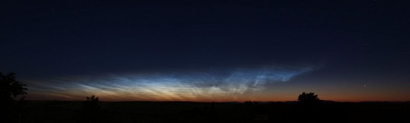 Wispy, glowing white clouds in dark sky over a black horizon with a dim sunset along it.
