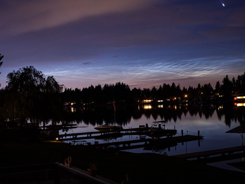 Lake with trees and light of houses at the background. There is a lake reflecting bright lights on a pink and dark blue sky.