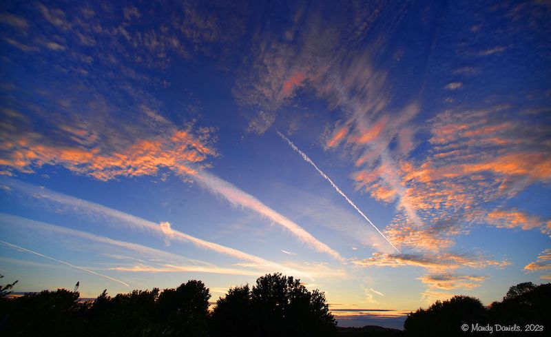 Golden-orangish clouds in a dark blue sky after sunset, with narrow white cloud strips (contrails) radiating from sunset.