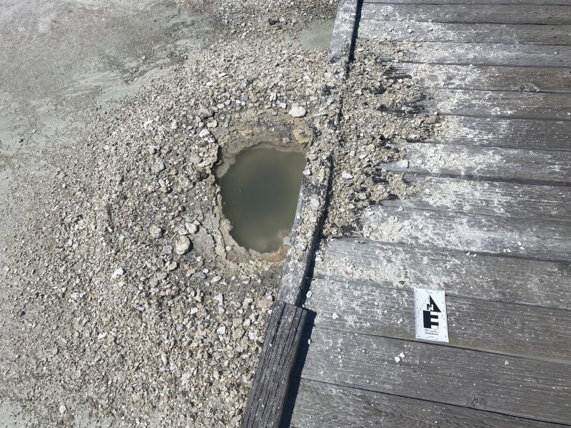 Yellowstone: A water-filled hole in the ground with gravel around it, and some debris thrown up onto a boardwalk.