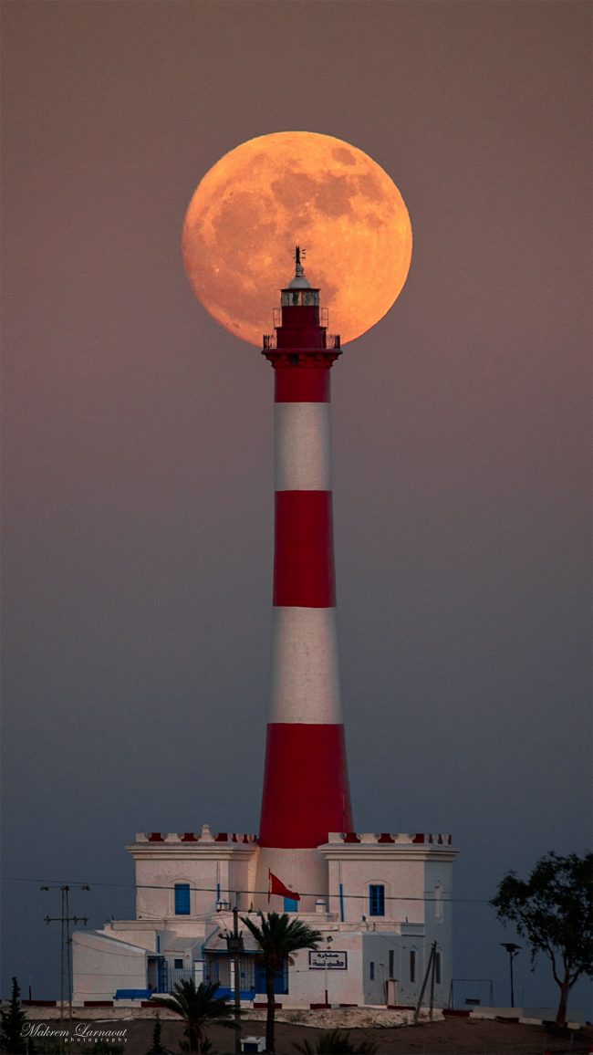 July supermoon: Tall red and white striped lighthouse with full moon behind the very top.