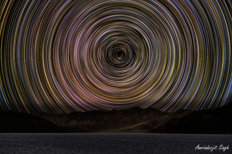 Above the silhouetted mountains, star trails glow in bright, colorful arcs circled around a central point in the sky.