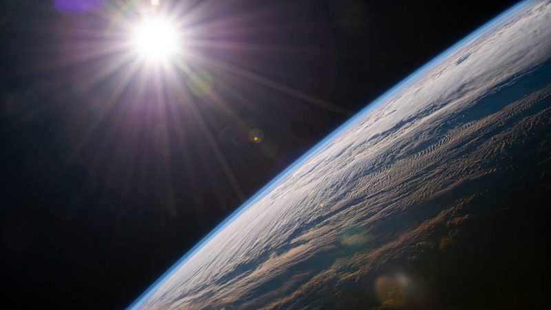 Day is 24 hours: Edge of Earth viewed from orbit with clouds and thin atmosphere while bright white sun shines in background.
