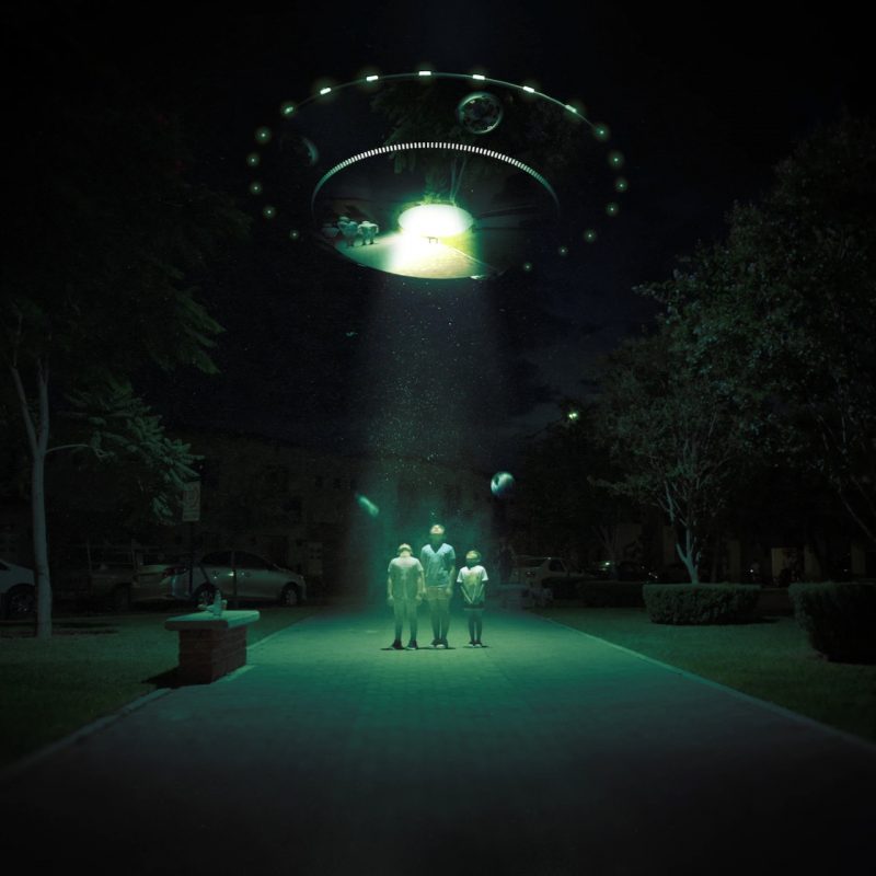 UFOs: Three people standing in a beam of light from a saucer shaped craft hovering above in darkness.
