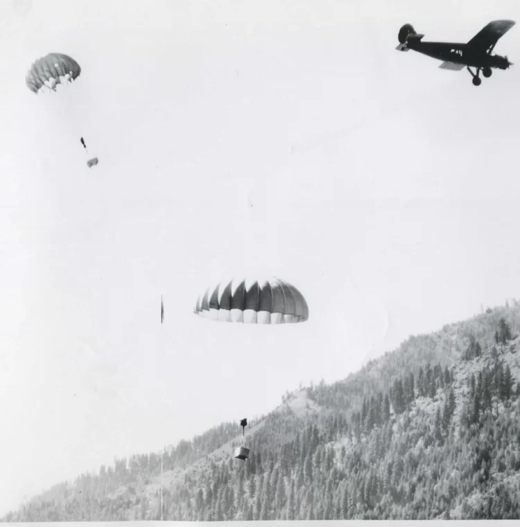 A small plane leaves the area as 2 parachutes holding wooden boxes fall toward a forested mountain.