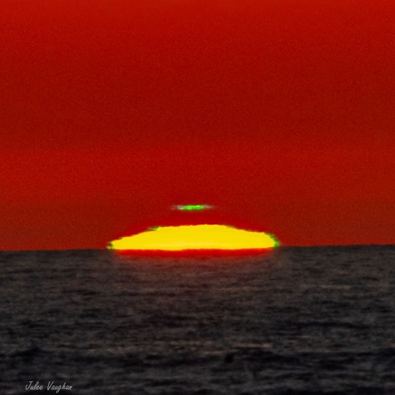 The glowing yellow top of the sun, on ocean horizon, with a short green streak floating above the sun.