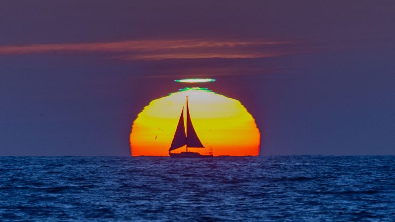 A boat with the sun right behind it. There is a green light at the top of the sun and some orange clouds.