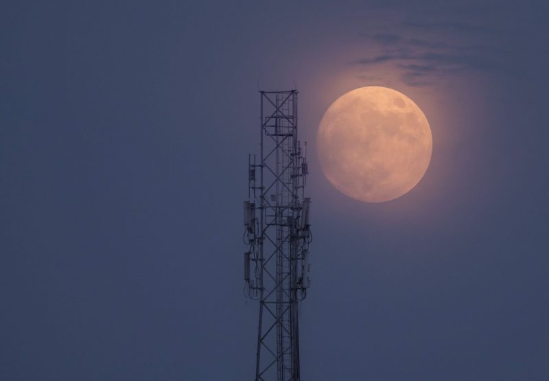A metal tower in the foreground with a hazy moon in the background.