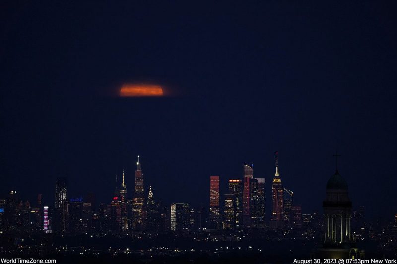 The middle part of a orange moon over very high buildings of different shapes and covered in lights.