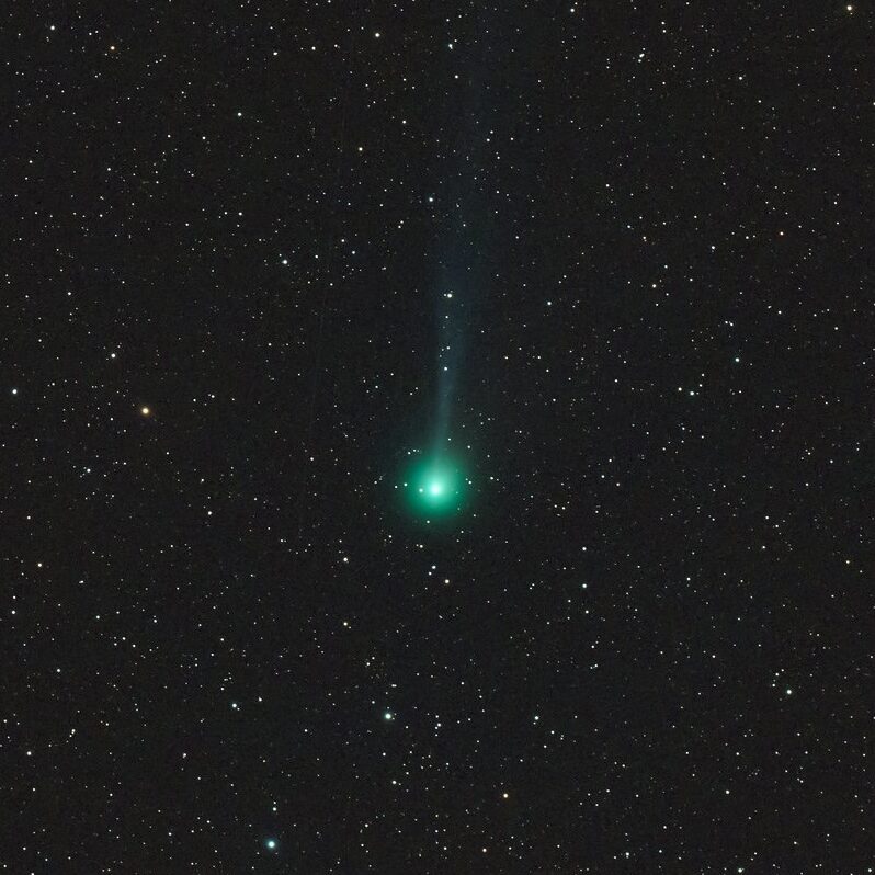 A bright greenish fuzzy ball with a tail leading straight up, in a star field.