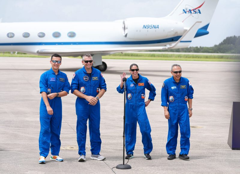 Four people in blue jumpsuits, with microphone, in front of a white plane with the NASA logo on its tail.