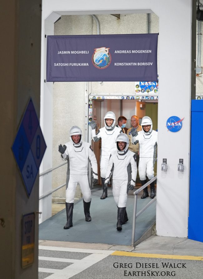 Crew-7 launch: four persons in space suits with helmets emerging from a building and waving.