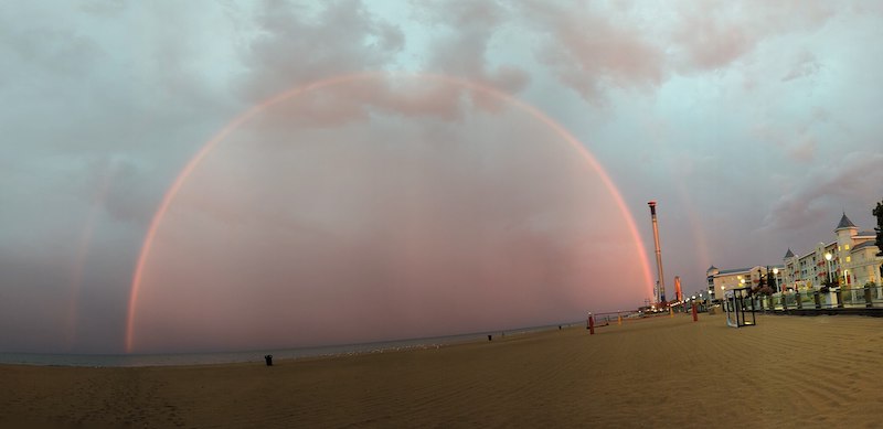 Beach with building on the side and a large pink double rainbow under cloudy sky over the ocean.