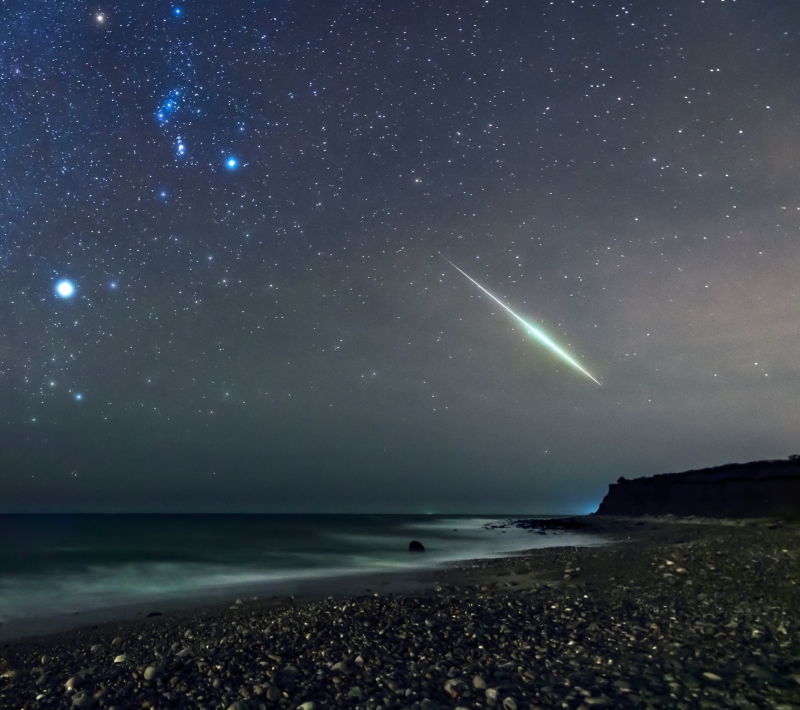 Long green line of a meteor above a beach, with constellation Orion and bright star Sirius below it.