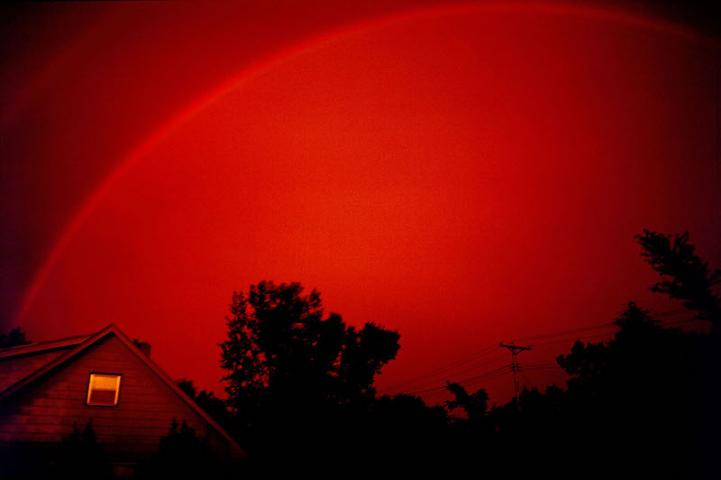 Entire sky ruby red, with a house and trees in foreground with part of a red rainbow arcing above.