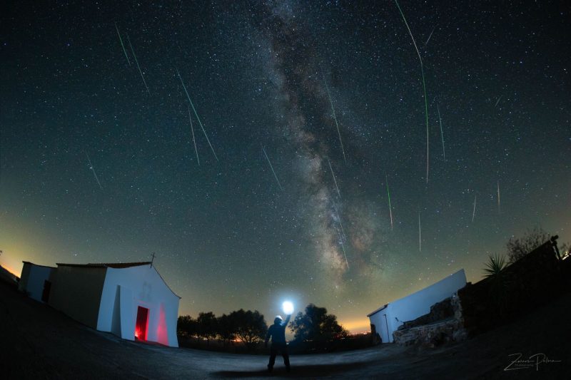 Many bright, thin meteor streaks falling from top of sky, with Milky Way nearby, over nearby buildings.
