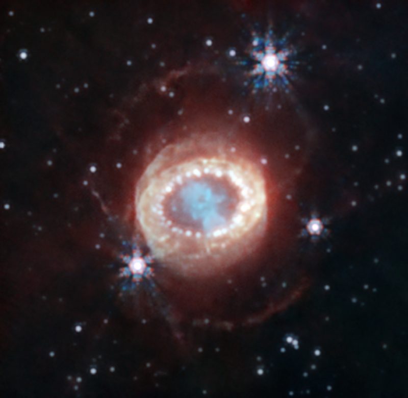 Supernova 1987A: In starry black sky, a wispy blue nebula surrounded by a ring of dots and concentric orangish layers.