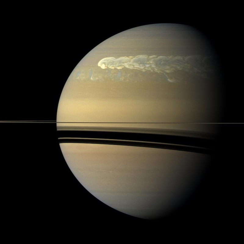 Storms on Saturn: Planet with large, long cloud in its northern hemisphere and thin ring seen edge-on casting a shadow beneath it.