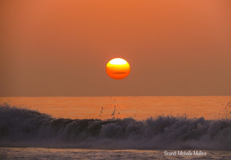 Orange sky and sea with waves in the foreground and a orange sun in the middle of the image.