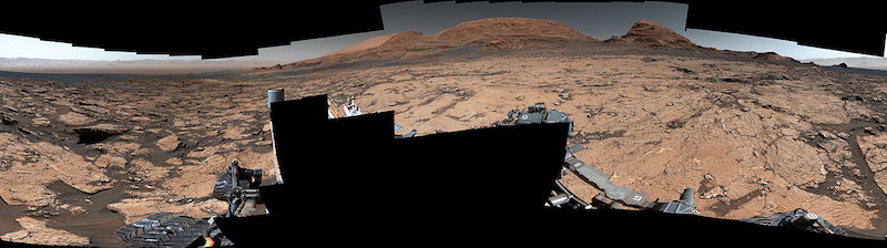 Wide view of rugged, reddish-brown Martian terrain with a distant hill.