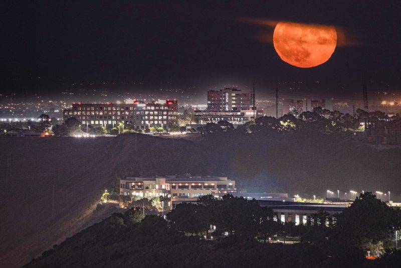 The bottom of a red full moon sticks out of clouds above a cityscape.