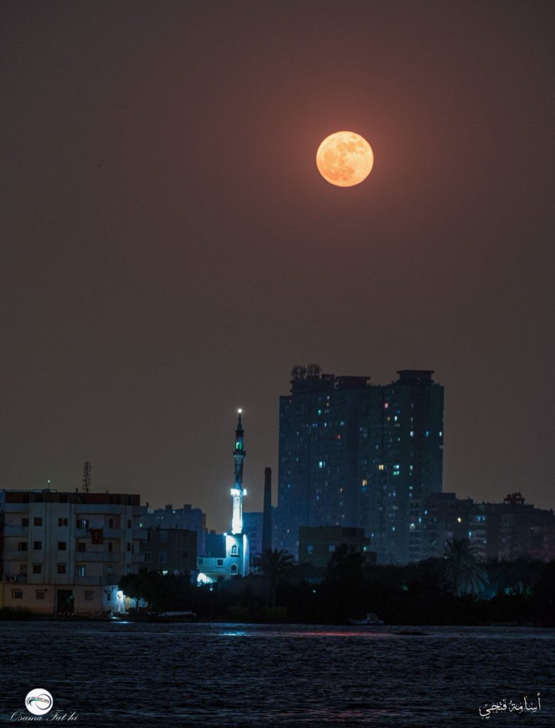 Glowing full moon over dark cityscape with a minaret.