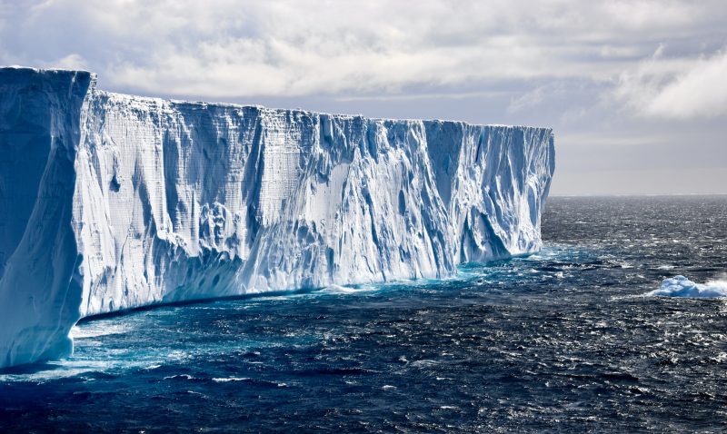 Antarctica: Sunny scene of a large iceberg with a flat top in the deep blue ocean with waves splashing around its base.