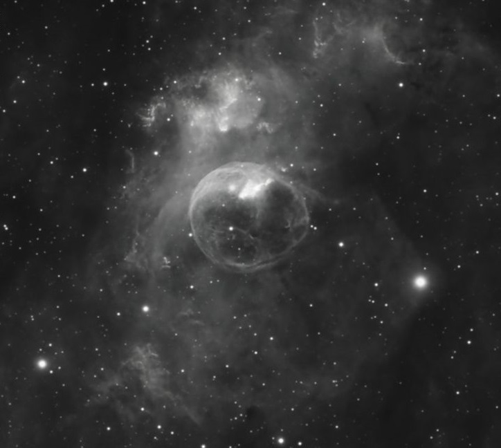 Black and white image of a translucent round bubble nebula inside dusty clouds of gas in a star field.