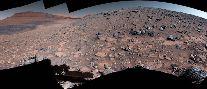 Curiosity rover: Brownish hill covered in sharp-edged rocks with larger layered hill in the background.
