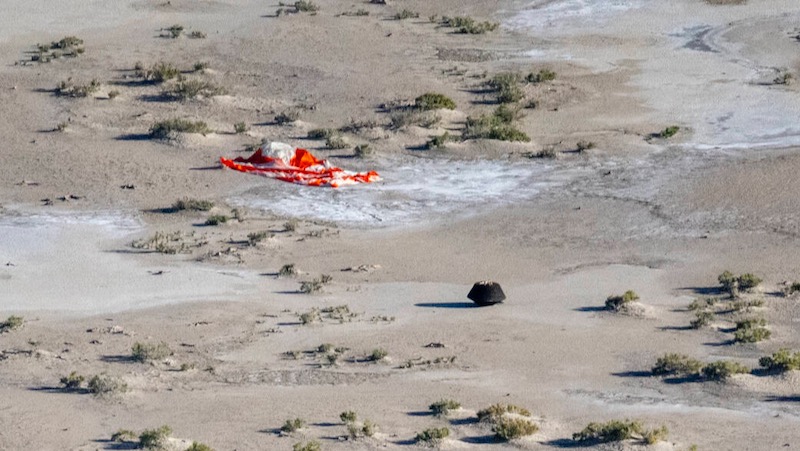 Sandy, tan desert, scattered scrubby bushes, small black spacecraft and red collapsed parachute on the ground.