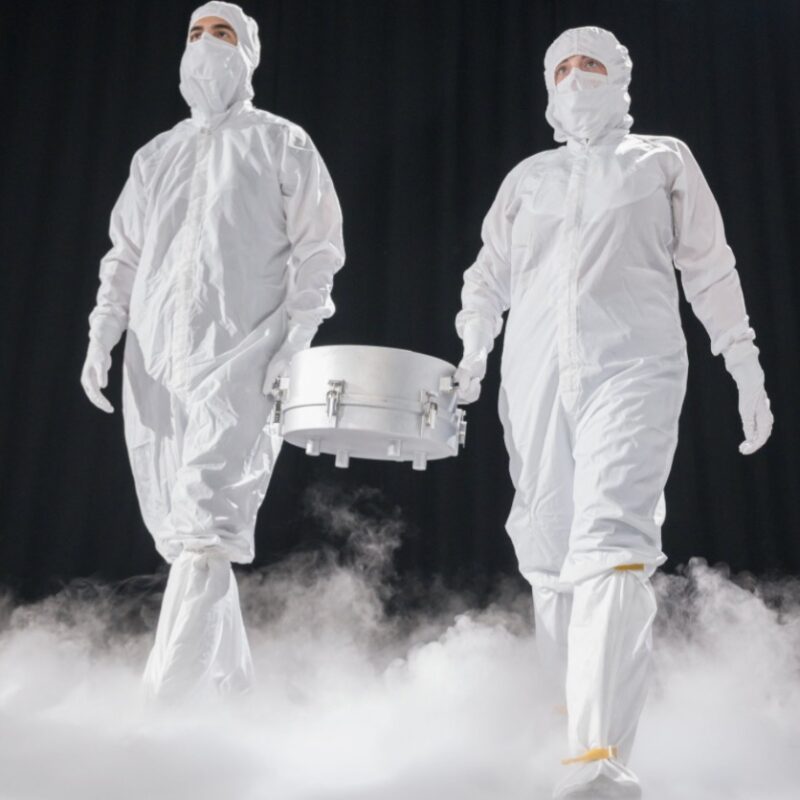 Two men in white suits carrying a silver canister between them, in a dark area with white mist boiling up from the floor.