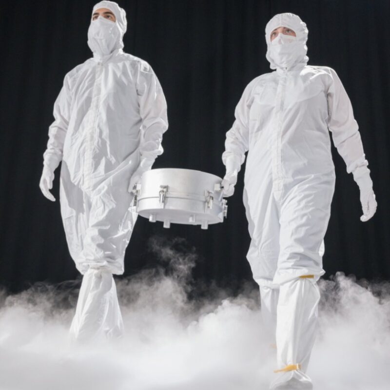 Two men in white clean suits carrying a silver canister, in a dark area with white mist boiling up from the floor.