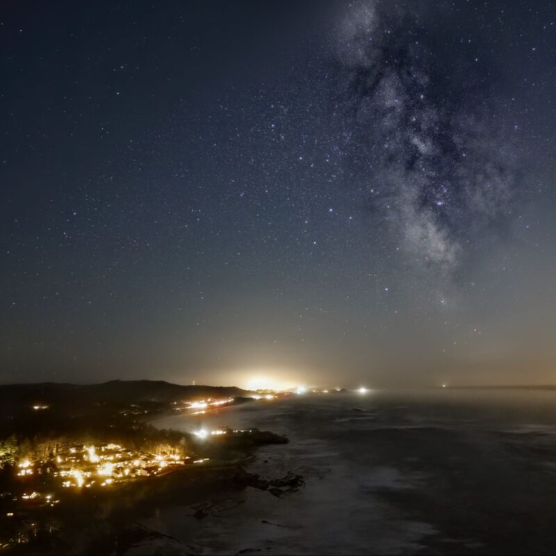 Noctalgia: Sea town with lights and more lights on the horizon plus Milky Way at upper left.