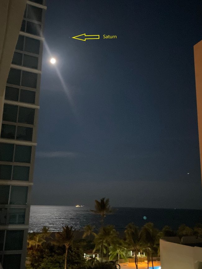 Bright moon beside a tall building, looking toward the ocean, with labeled Saturn above.