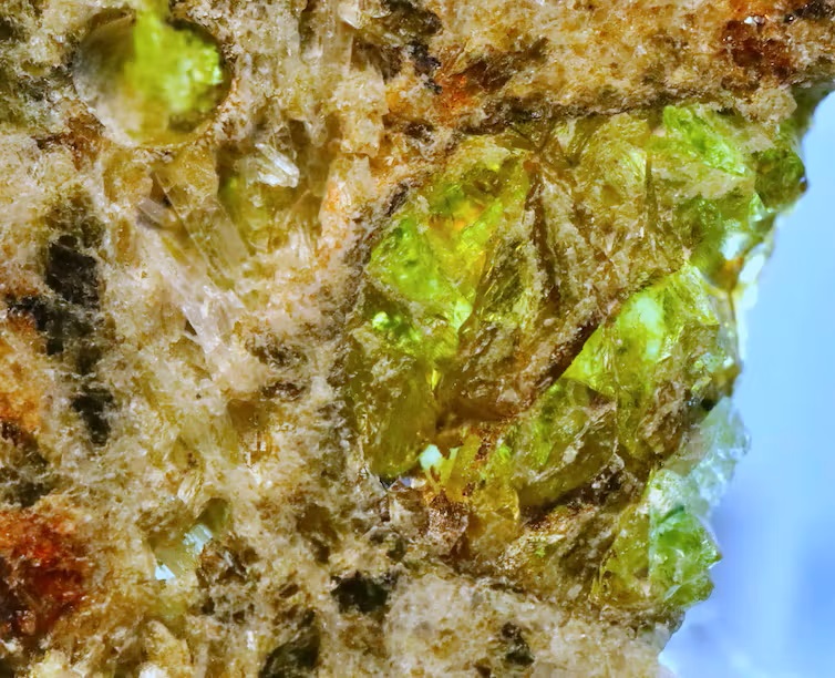 Irregular tan space rock shown closeup with green, translucent crystals embedded in it.
